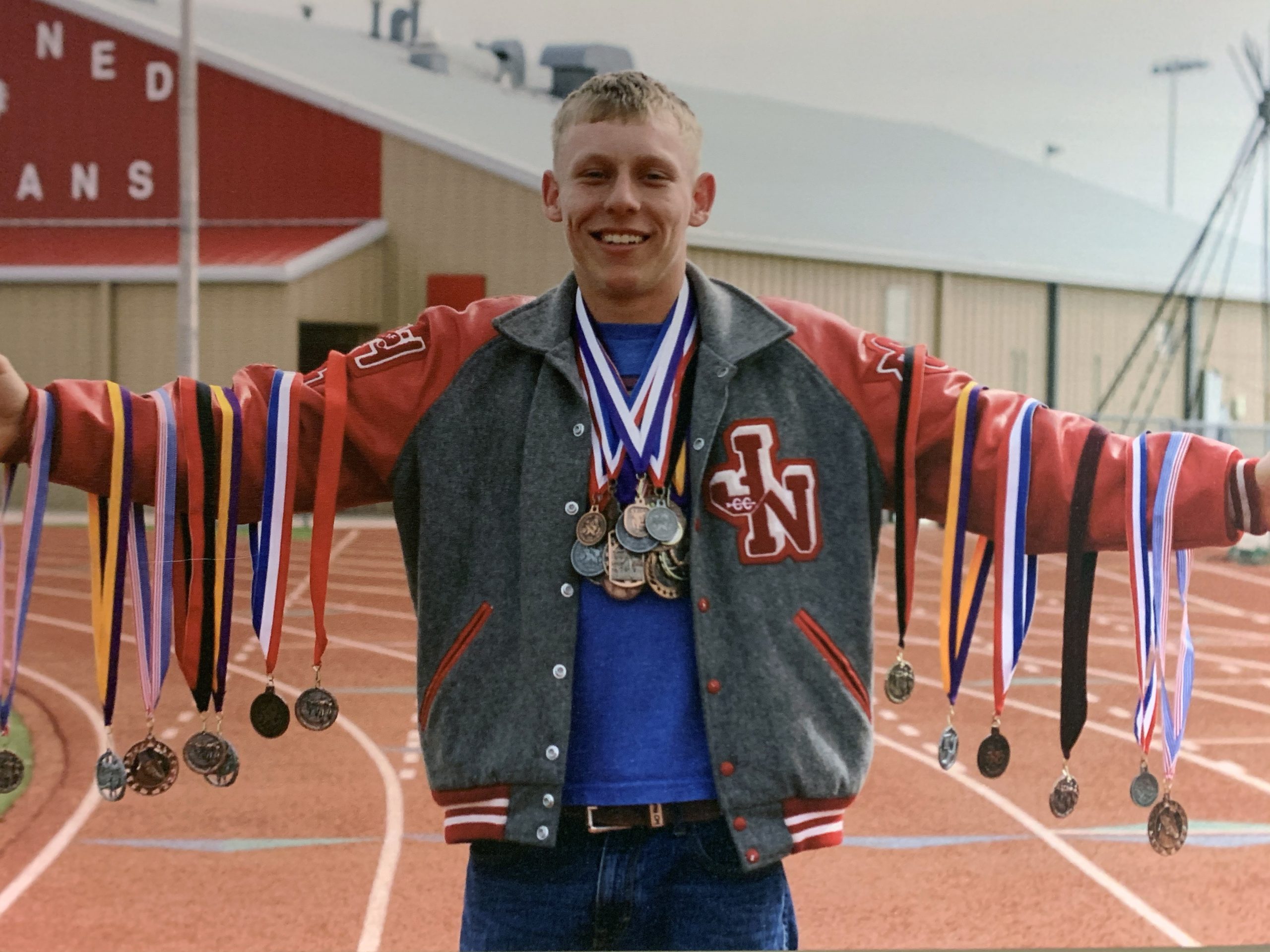 Young man with athletic jacket on with arms out holding athletic medals