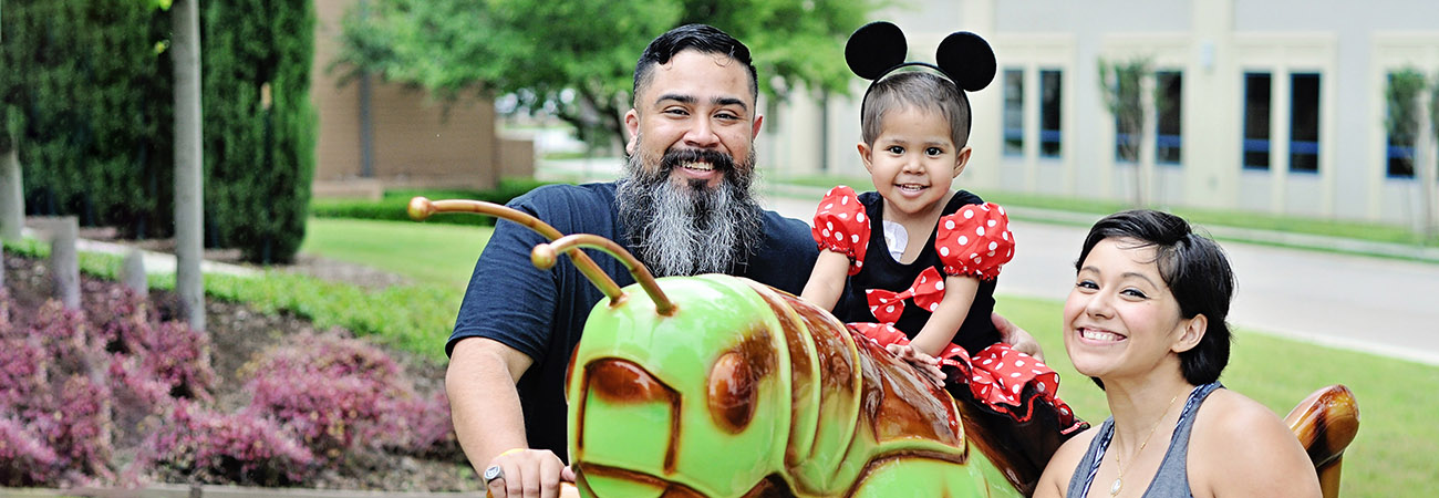 Mom and dad smiling at camera with little girl in minnie mouse ears sitting on a grasshopper
